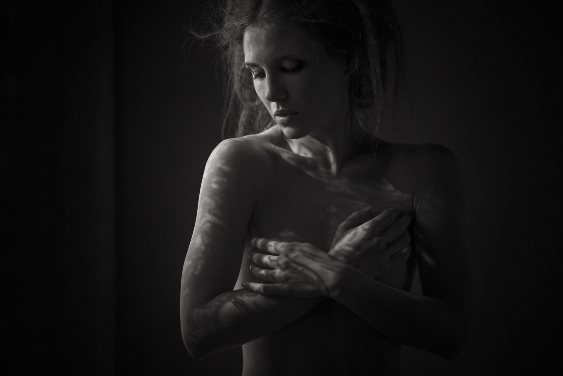 Woman with crossed arms over bare chest, head bowed, lit from side against black background in dramatic chiaroscuro style