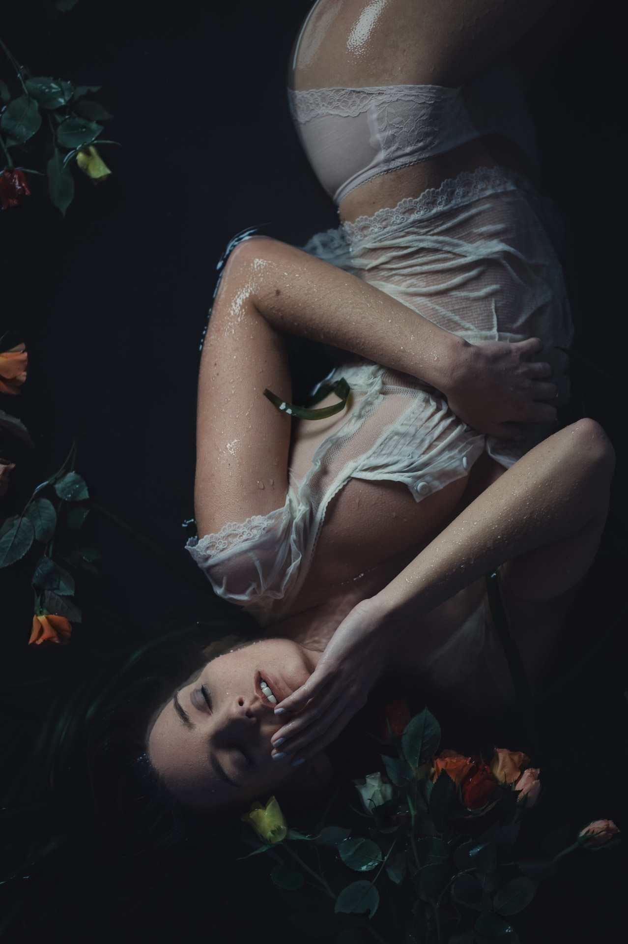 Woman floating in dark water wrapped in white fabric, surrounded by orange and green flowers, shot from above