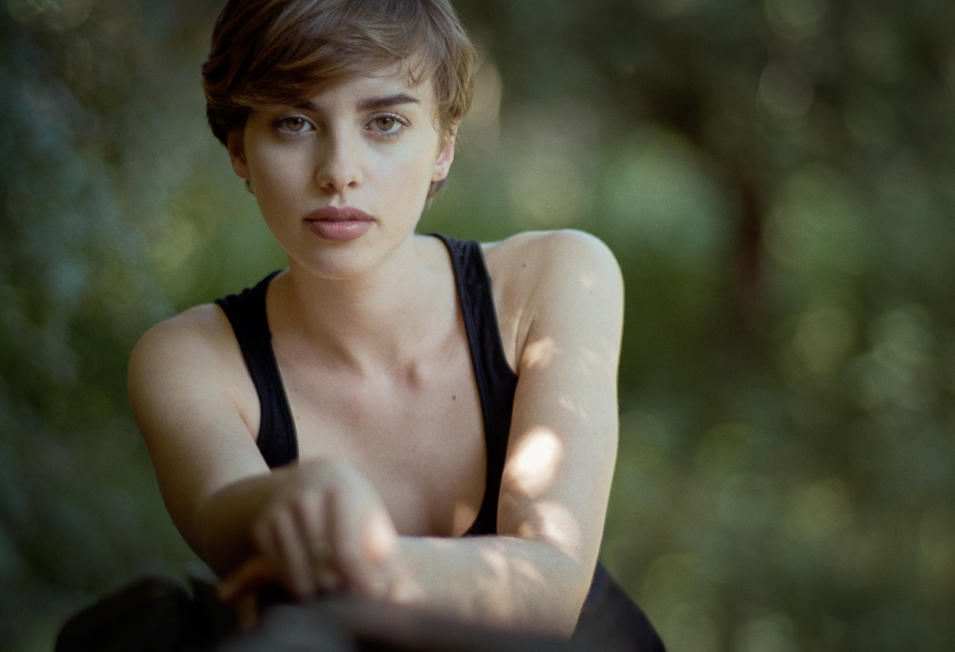 Woman with short blonde hair in black sleeveless top touches her arm, looking at camera with blurred green forest behind her