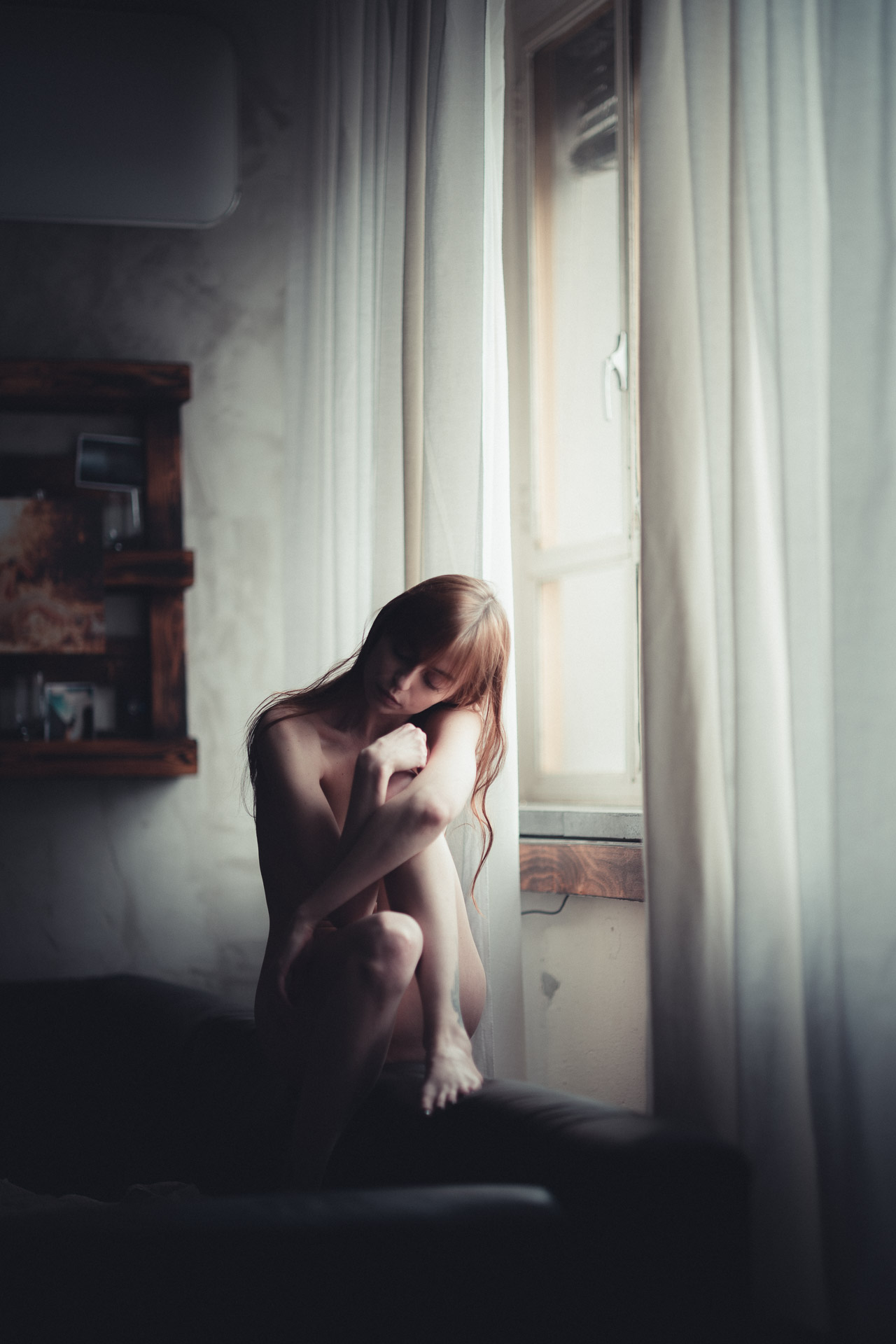 Nude woman in fetal position on windowsill, head bowed, surrounded by white curtains with natural light filtering through