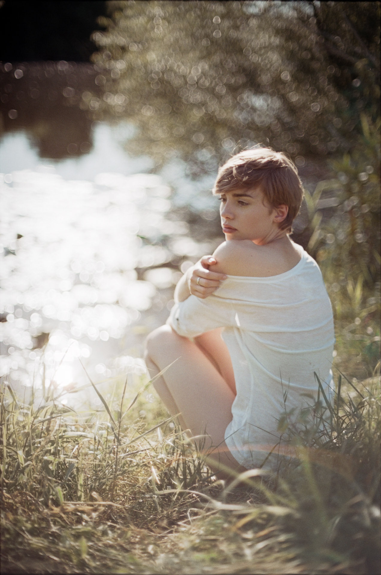 Young woman sitting by water wrapped in white fabric, looking pensively to the side with bokeh reflections in background