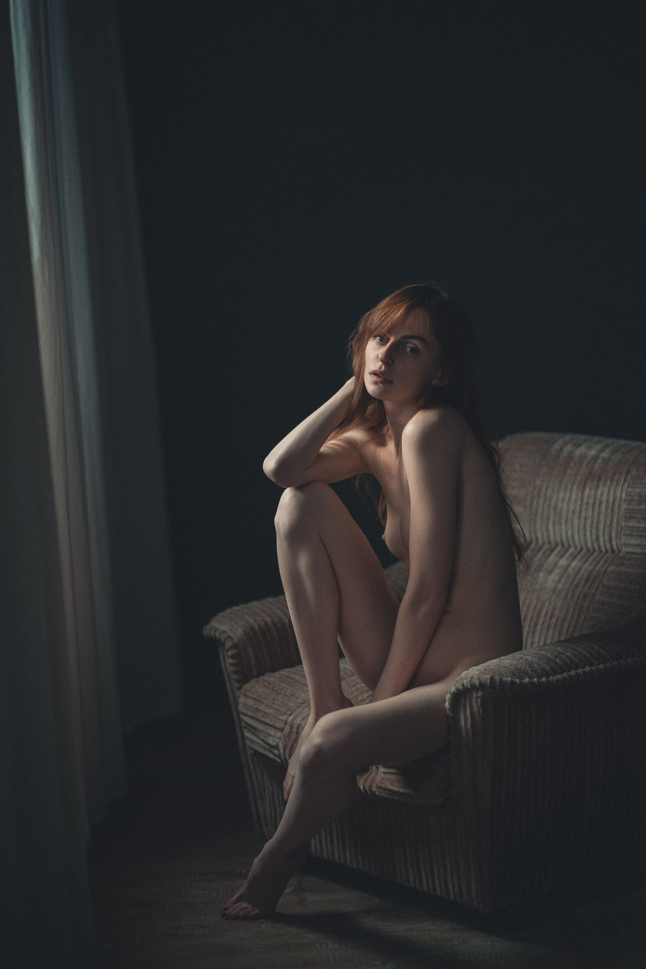 Woman with red hair seated on wicker chair in darkened room, knees drawn up, lit by soft window light from left side
