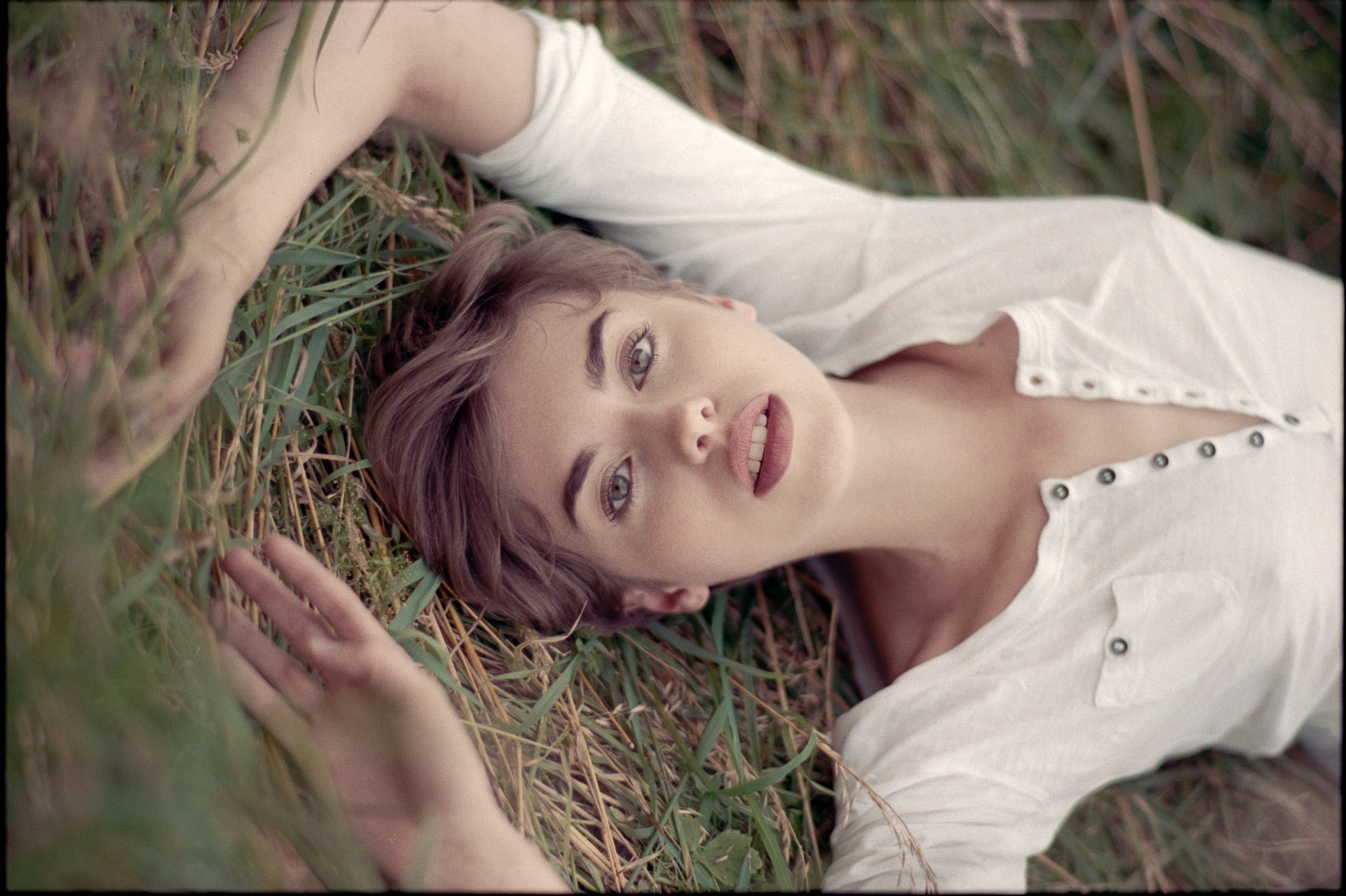 Woman with purple hair lying on grass, wearing white studded top, photographed from directly above as she looks up at camera