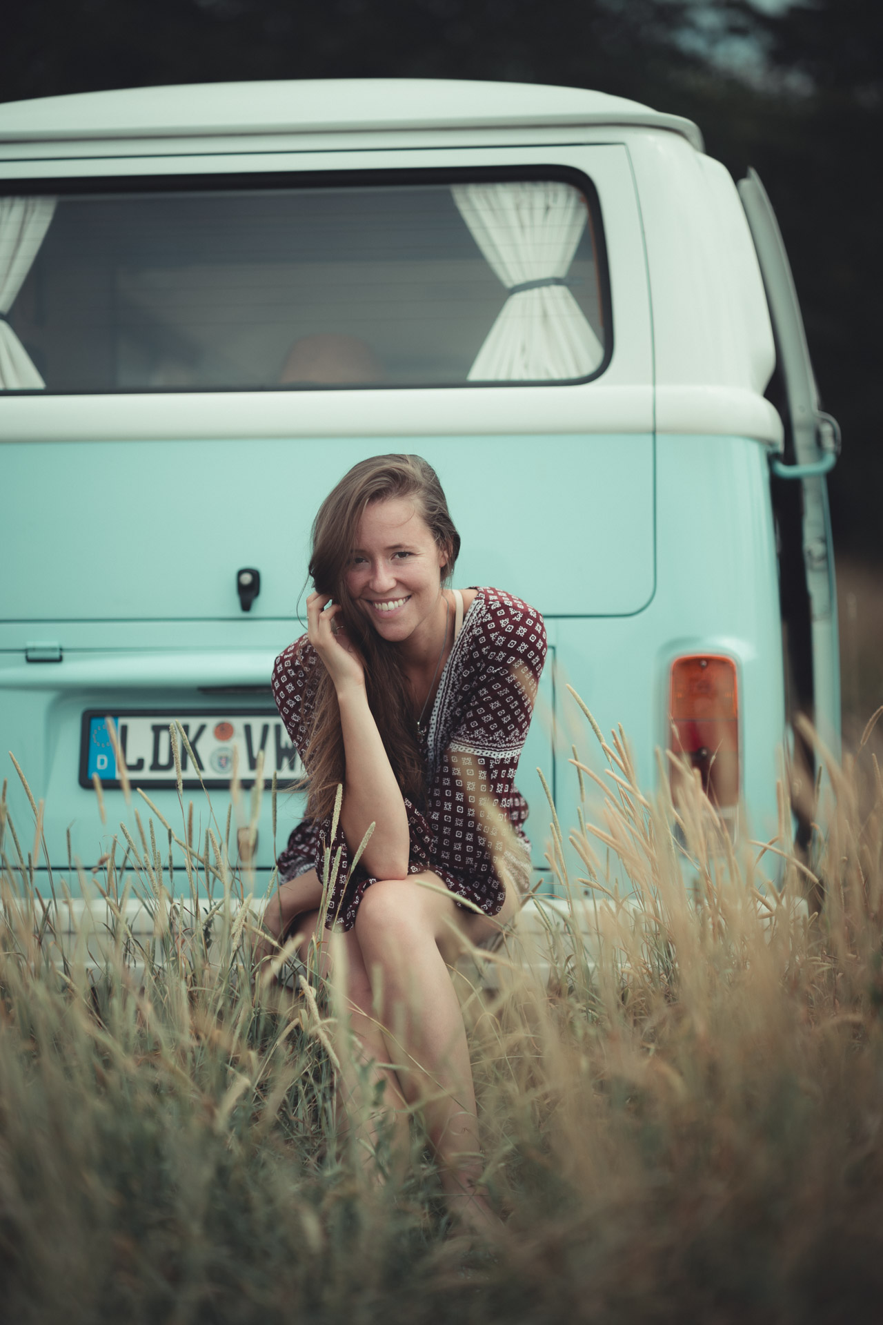 Woman in polka dot dress smiling, seated in tall grass in front of mint green VW van with license plate reading ALDK*VN