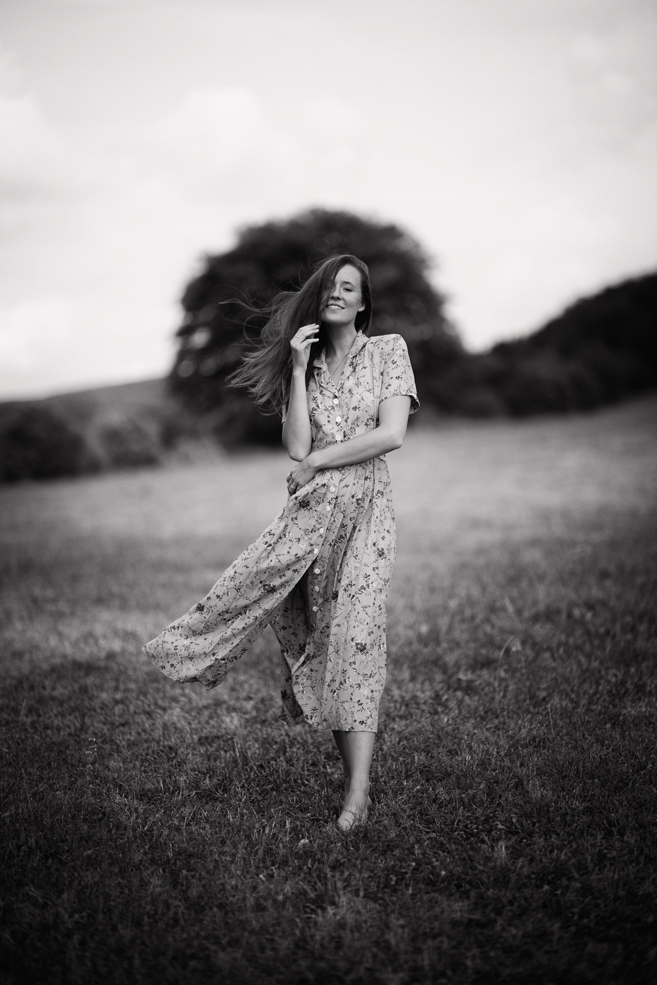 Woman in floral dress standing in windswept meadow at dusk, hair blowing dramatically, hand touching face, trees in background