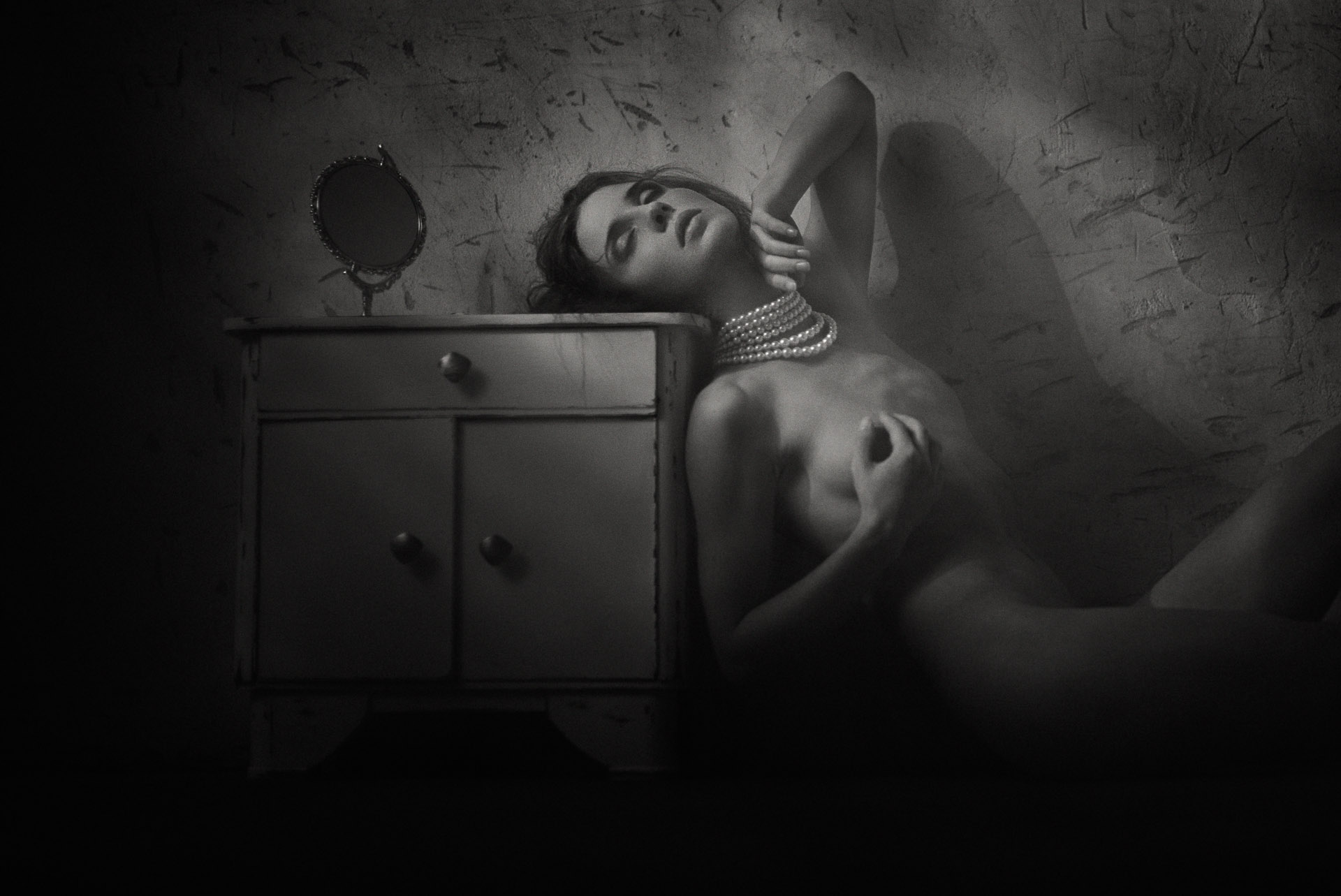 Woman lying on floor beside vintage nightstand with mirror, head tilted back, hand behind head, shot from above in black and white