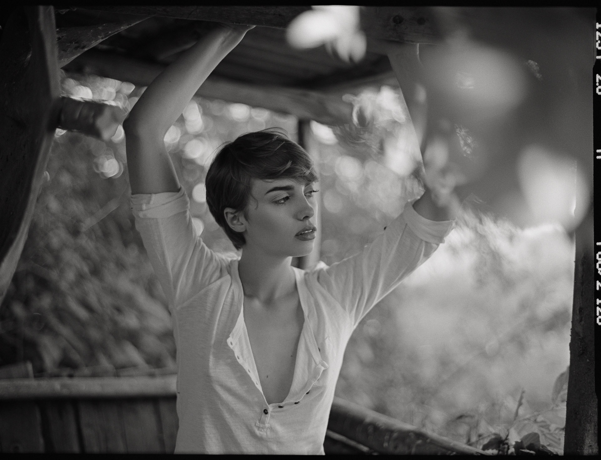 Woman with short dark hair in white unbuttoned shirt, arms raised above head, standing outdoors with blurred bokeh background