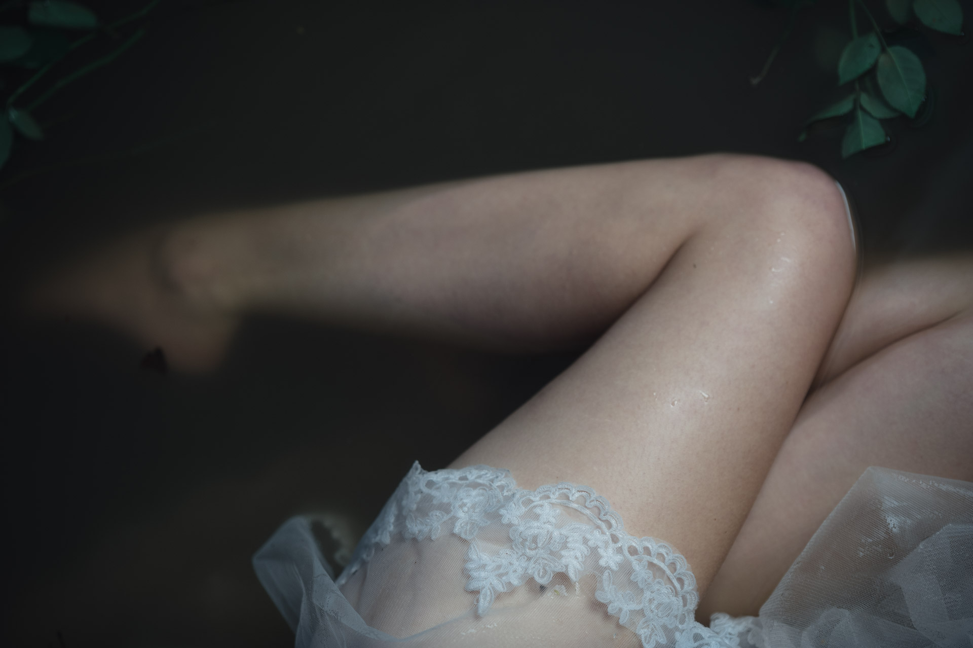 Bare legs and thighs with white lace and tulle fabric gathered at hip level, photographed against dark background with plant leaves visible