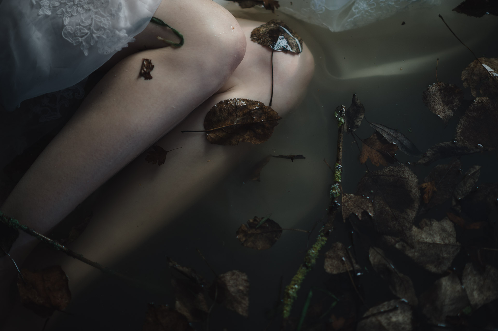 Pale arm resting in shallow water among moss-covered rocks and debris, with a small key tattoo visible on the wrist