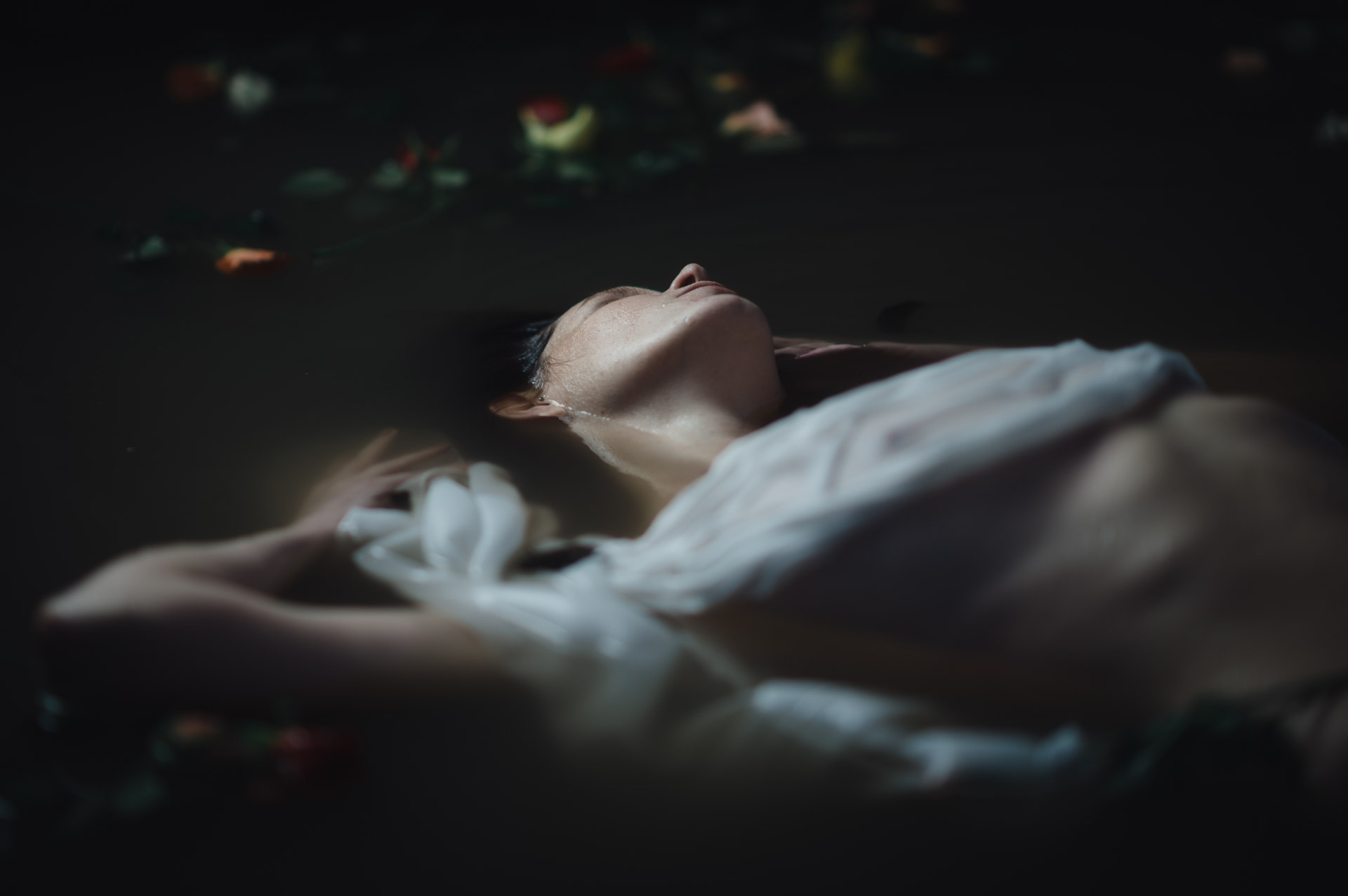 Woman lying back with closed eyes, head tilted, loose hair falling, bare shoulder visible, flower petals floating above in darkness