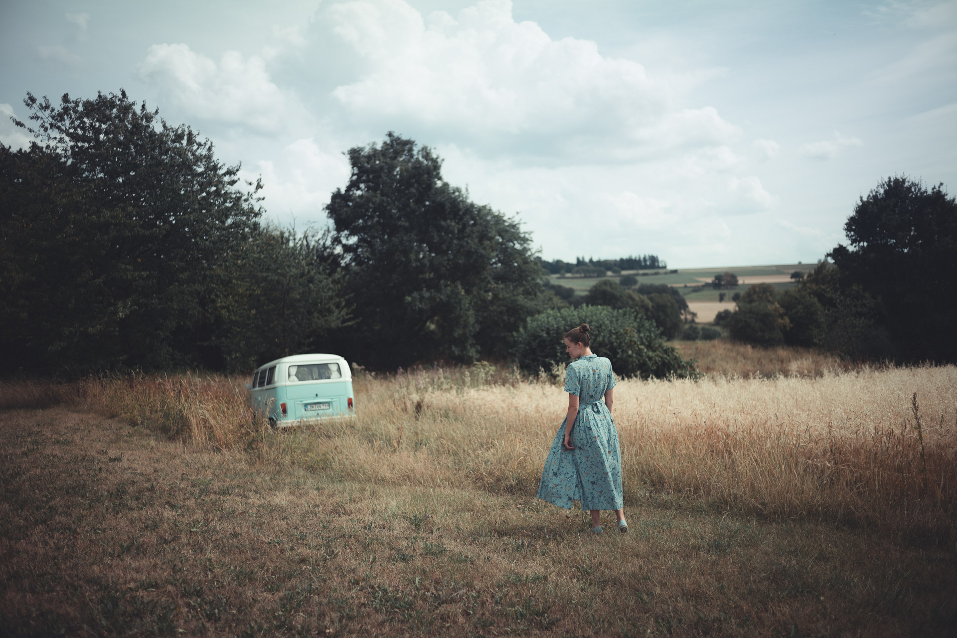 Woman in long dress stands in dry grass field next to vintage blue caravan, with trees and overcast sky in background