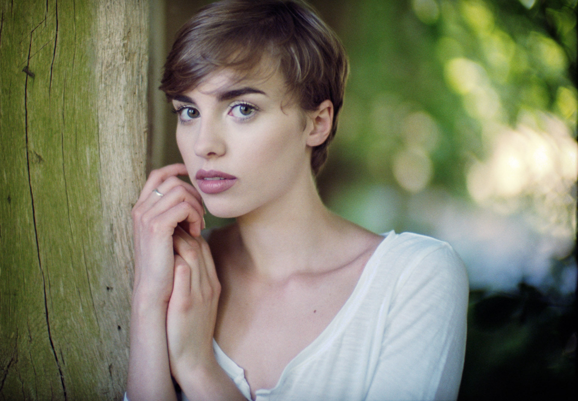 Woman with short brown hair in white top leaning against moss-covered tree, hand near face, looking at camera in outdoor setting