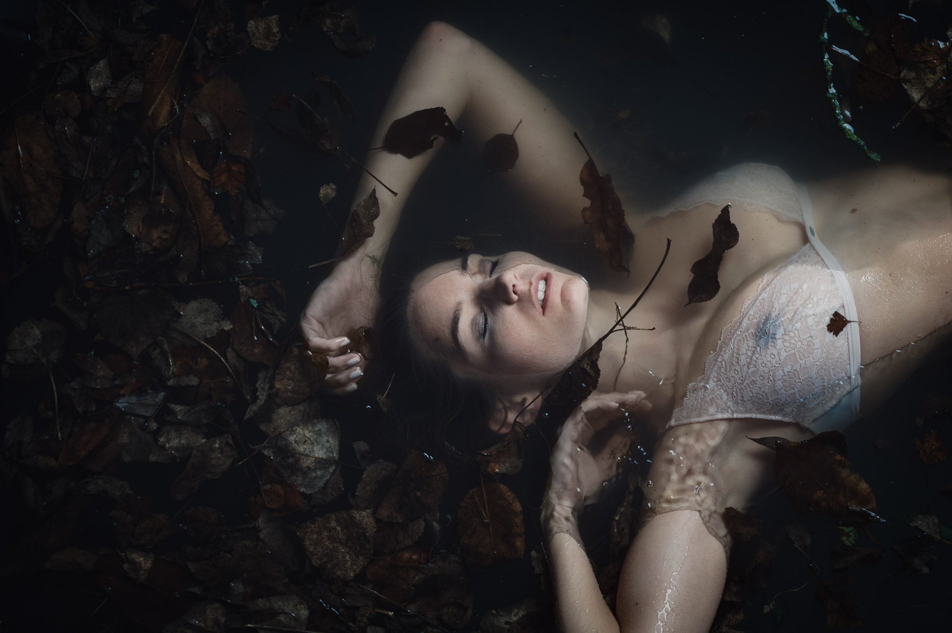 Woman lying on back among autumn leaves, arms raised above head, wearing white undergarment, photographed from directly overhead