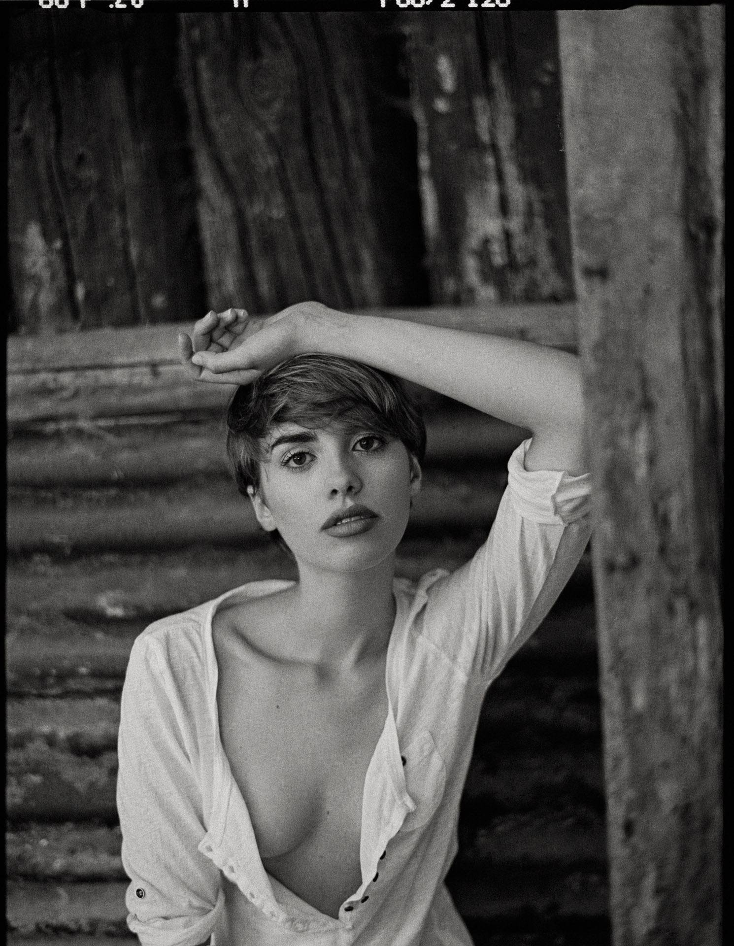 Woman with short dark hair in white open shirt, hand to forehead, against weathered wooden wall background