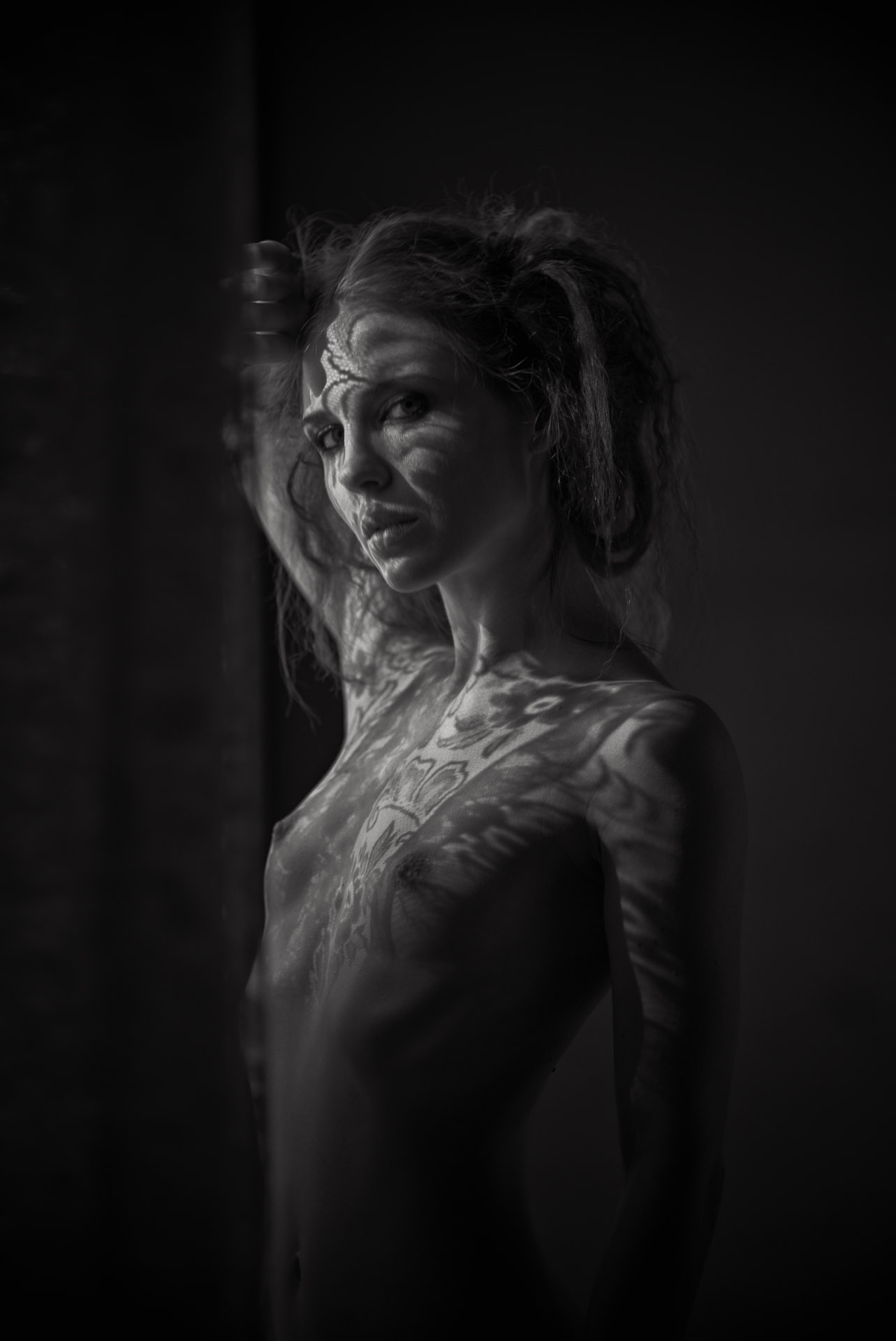 Woman with hair pulled back, photographed from chest up in profile against dark background, dramatic side lighting across bare skin