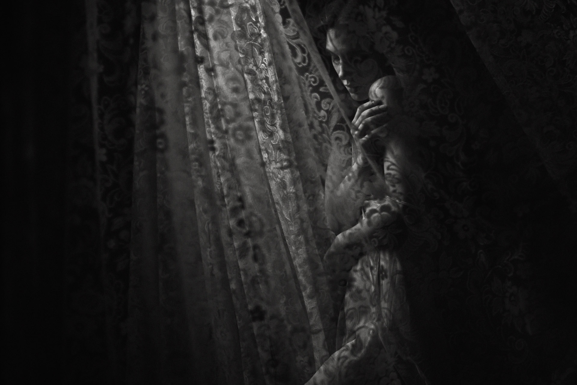 Woman in profile standing against ornate weathered curtain or tapestry with decorative patterns, dramatic side lighting
