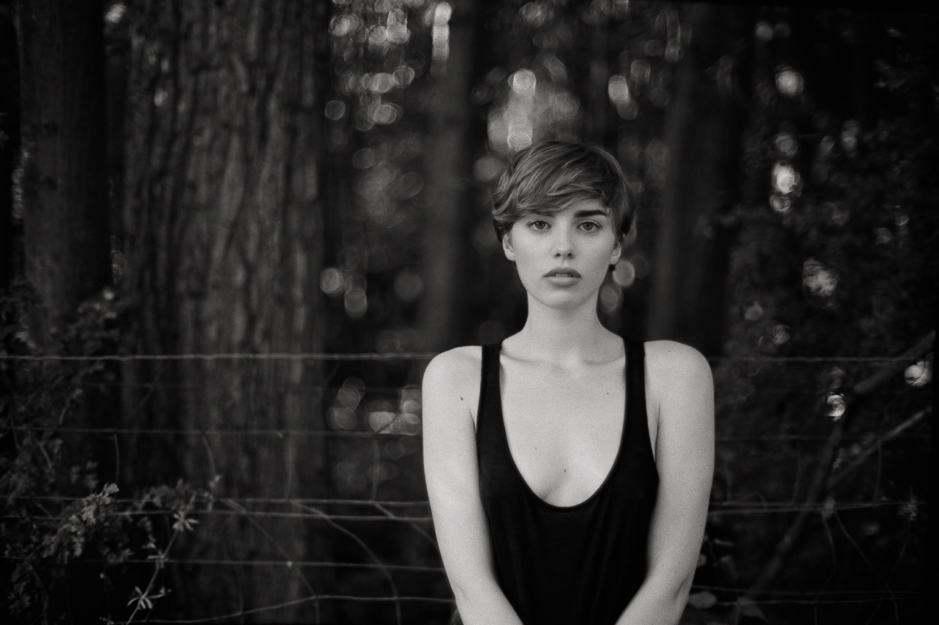 Young woman with short dark hair and bangs in black tank top, standing before blurred forest with barbed wire fence visible