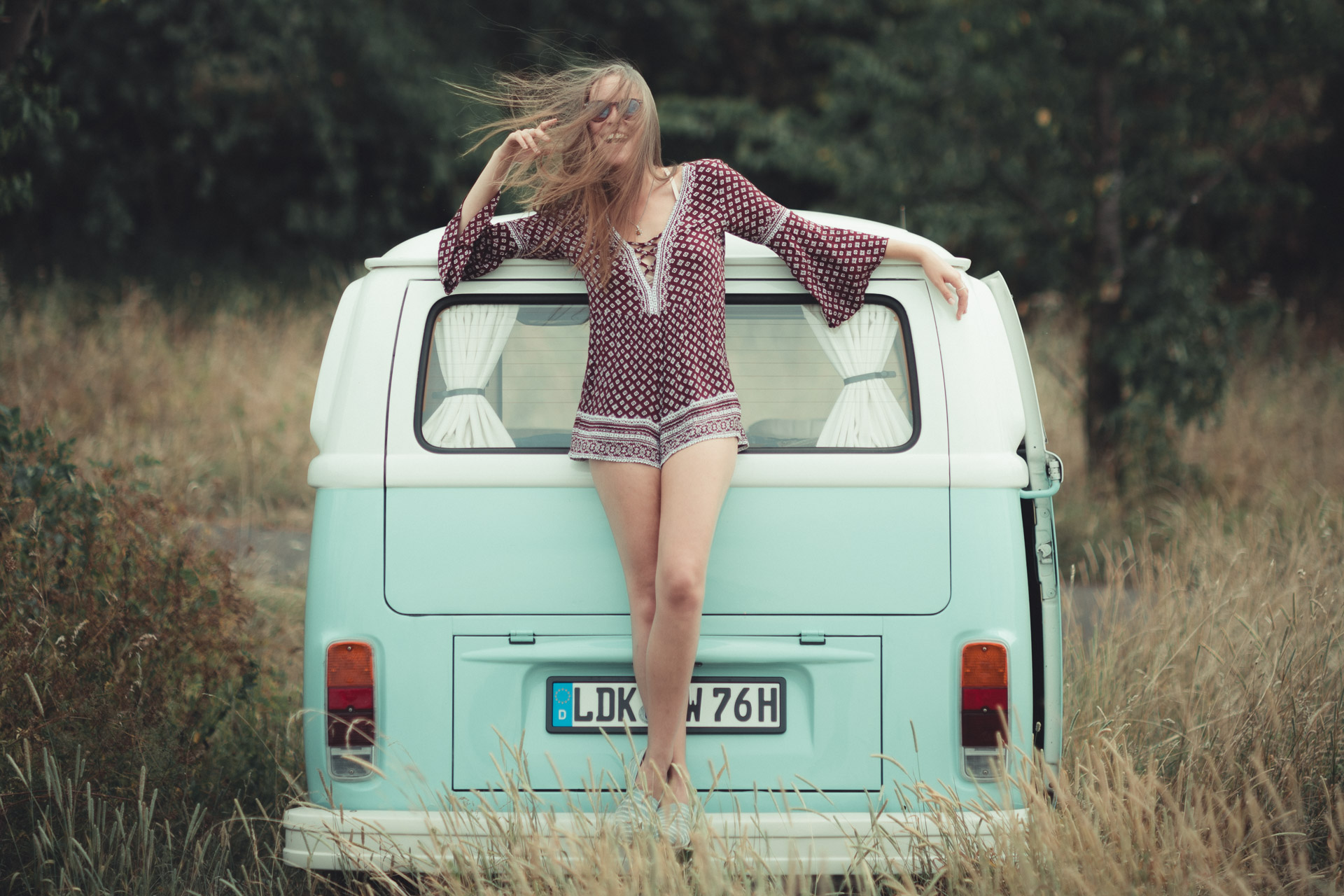 Woman in patterned top leaning back against turquoise vintage VW van in tall grass field