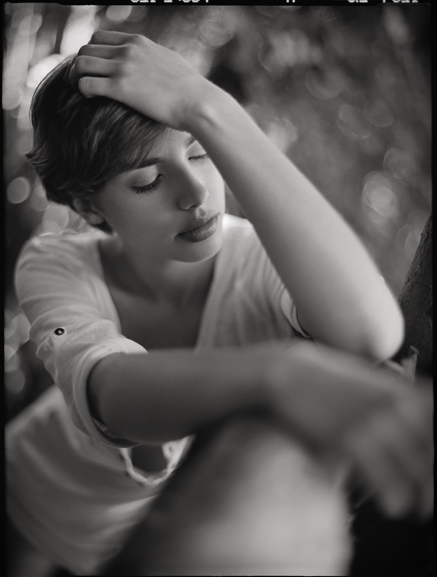 Young woman with short hair, eyes closed, hand on forehead in contemplative pose, wearing white t-shirt, shot from slight angle