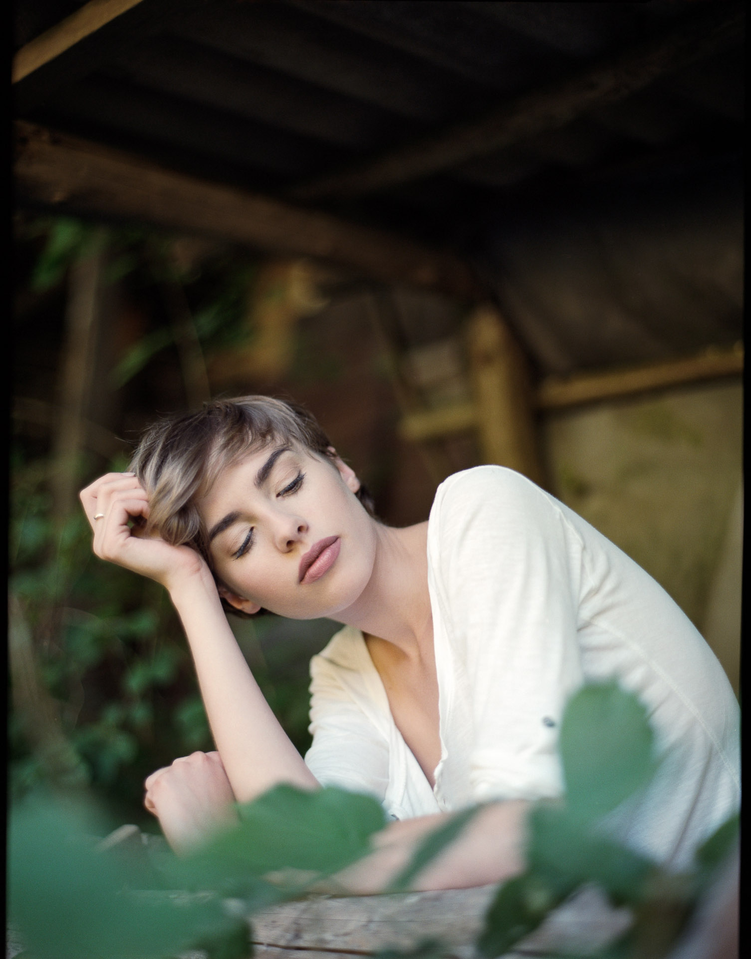 Woman with short brown hair in white blouse rests head on hand at wooden table, eyes closed, green foliage in foreground and background