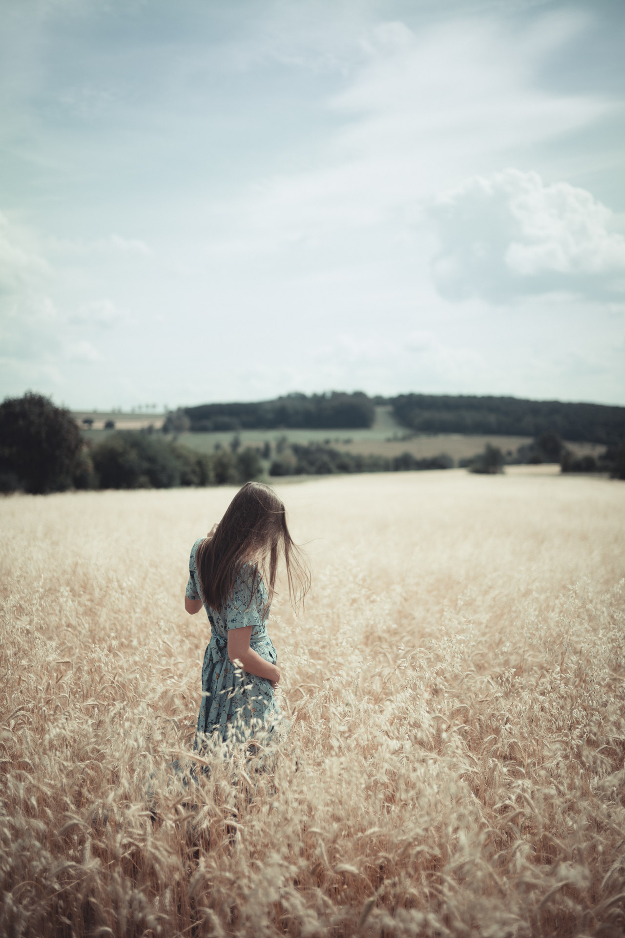 Woman with long brown hair in blue floral top and white shorts walking through golden wheat field toward distant trees