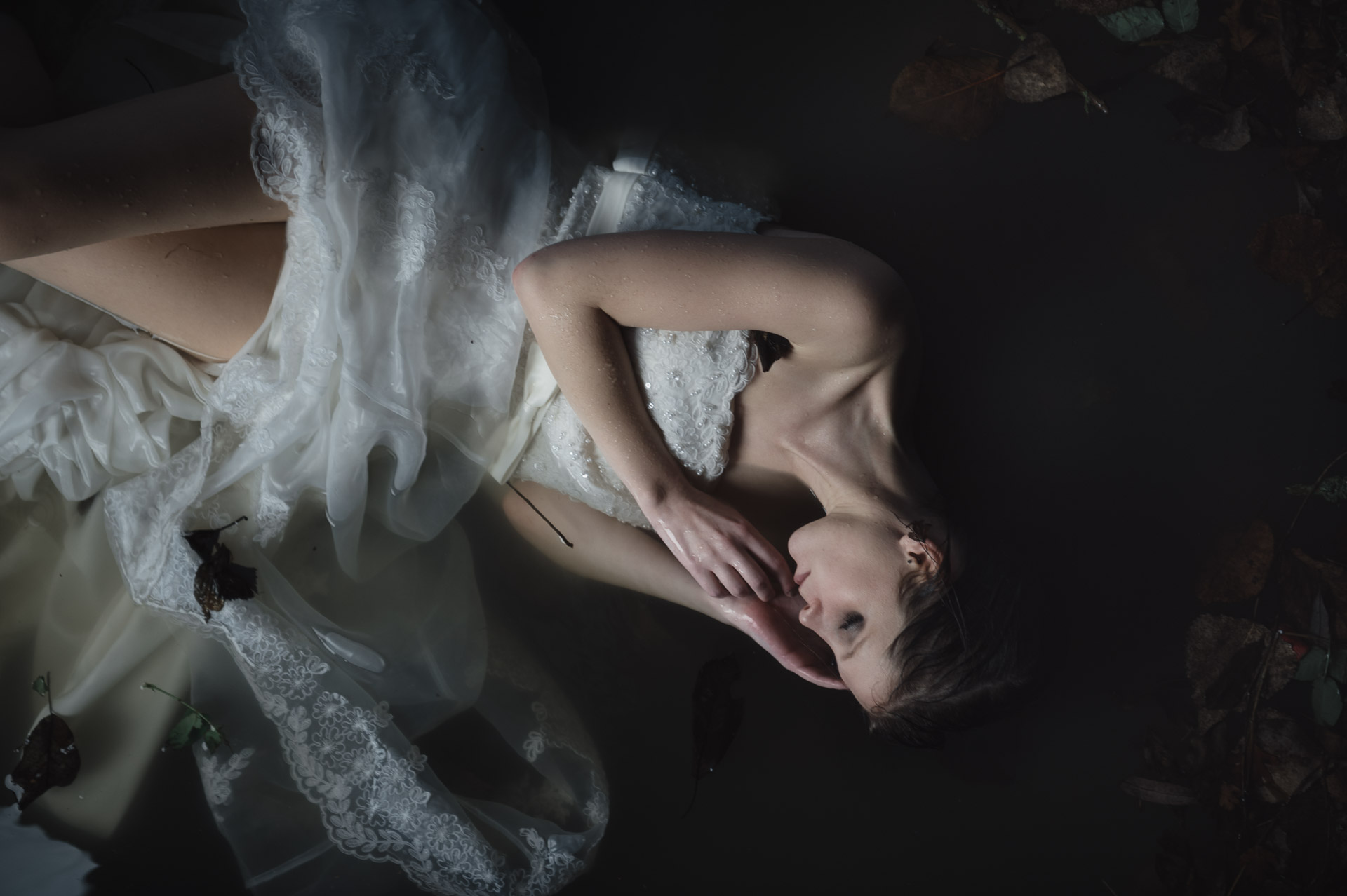 Woman lying on back with platinum hair spread out, wearing white lace dress, photographed from above against dark background