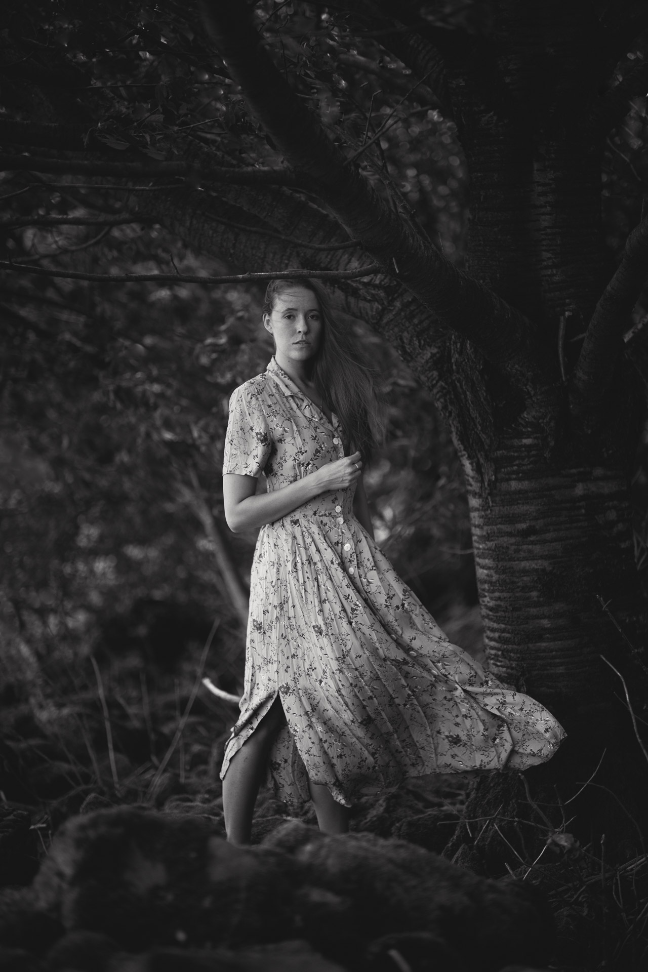 Woman in floral wrap dress standing in overgrown tropical garden with palm fronds and dense foliage, dress flowing at knee-length