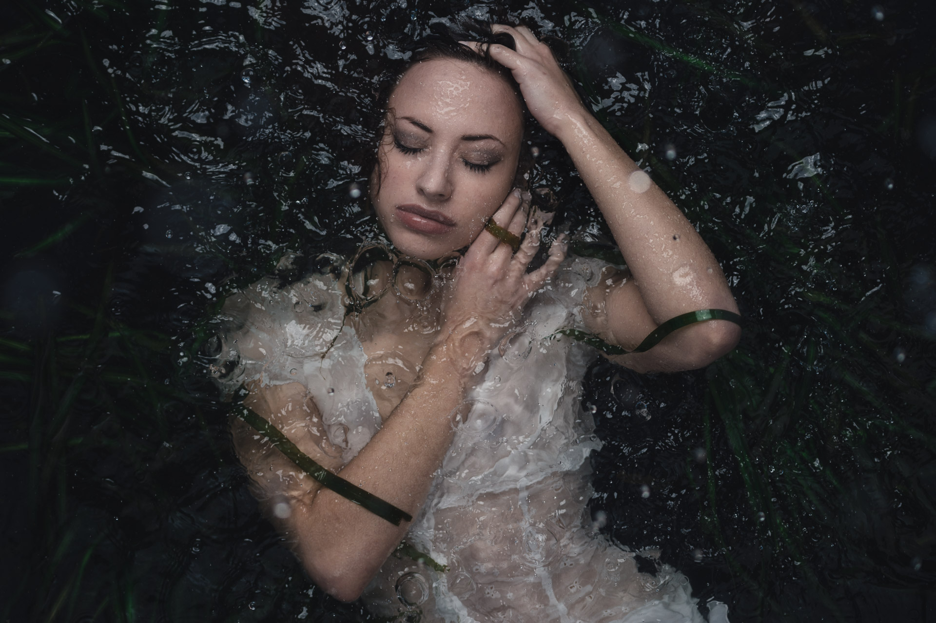 Woman submerged in dark water wearing white dress, face tilted back with hand near head, bubbles and particles floating around her