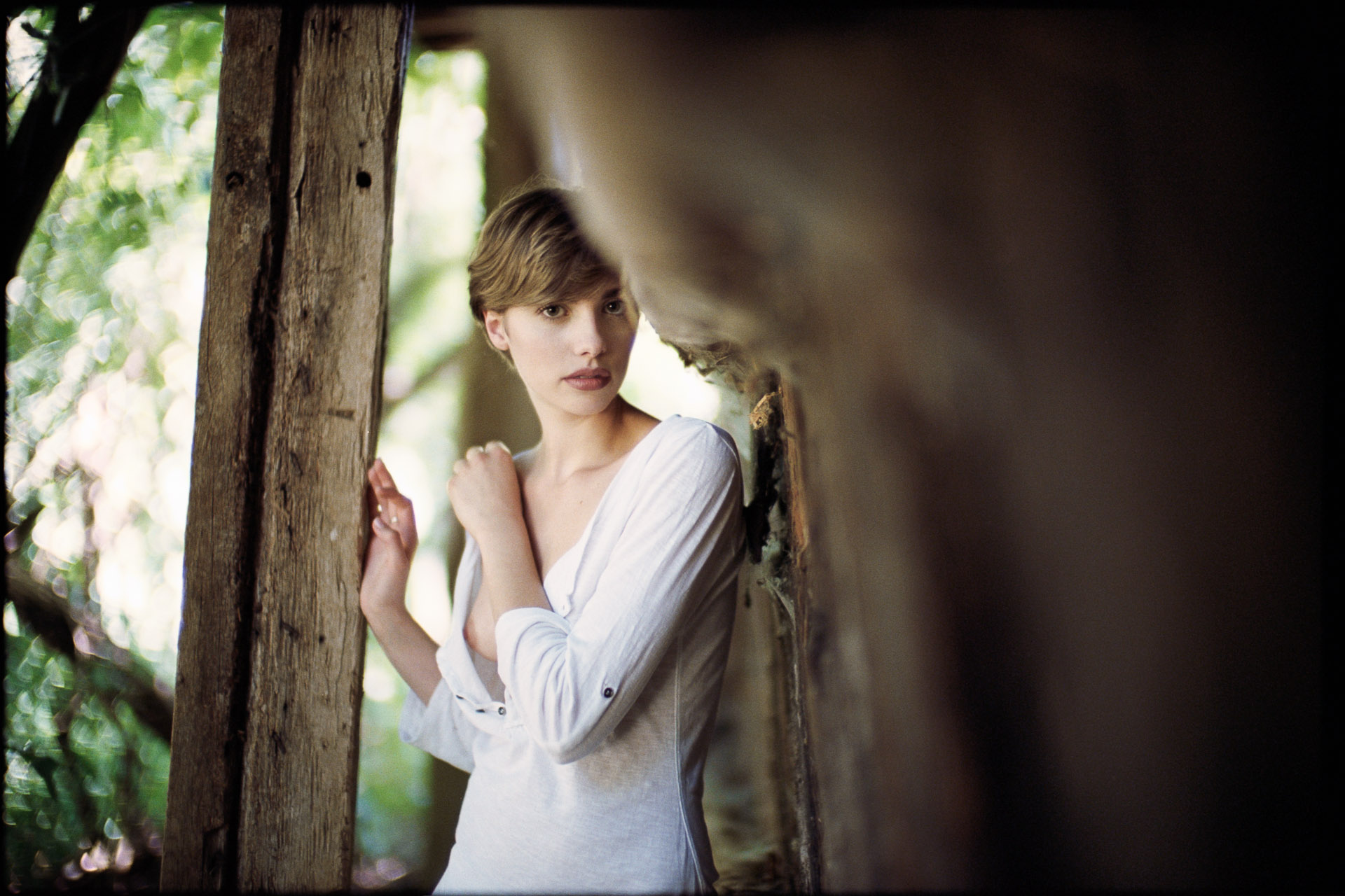 Woman in white clothing leaning against wooden beam in rustic interior with natural backlighting through doorway or window