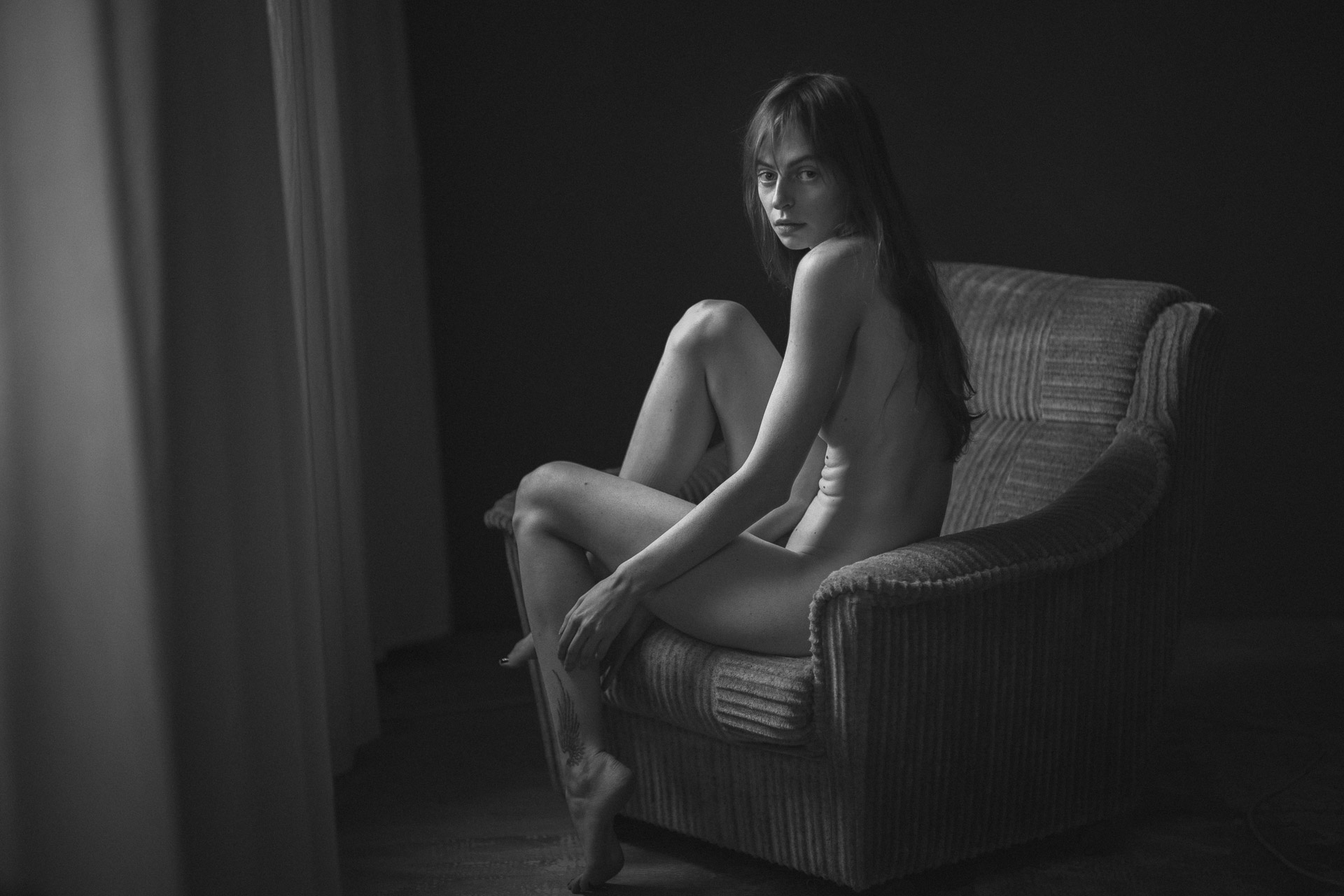 Nude woman seated in profile on textured armchair near window, arms wrapped around knees, head turned toward camera