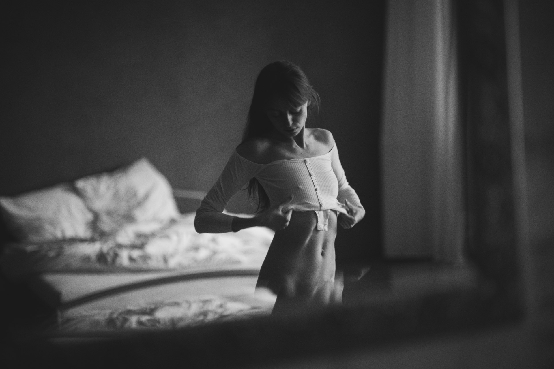 Woman in white lingerie standing beside unmade bed in dimly lit bedroom, backlit by window creating silhouette effect